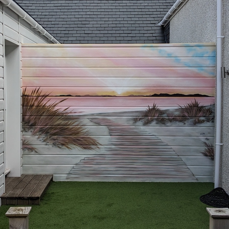 Mural painted on horizontal white siding depicting a boardwalk through sandy dunes toward a pink sunset over the ocean and distant hills.
