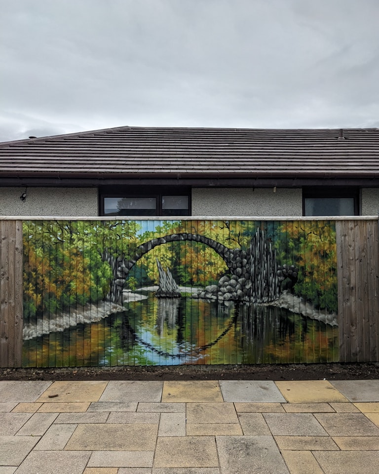 Mural depicting the Rakotzbrücke Devil's Bridge over water, painted on a wooden garden fence