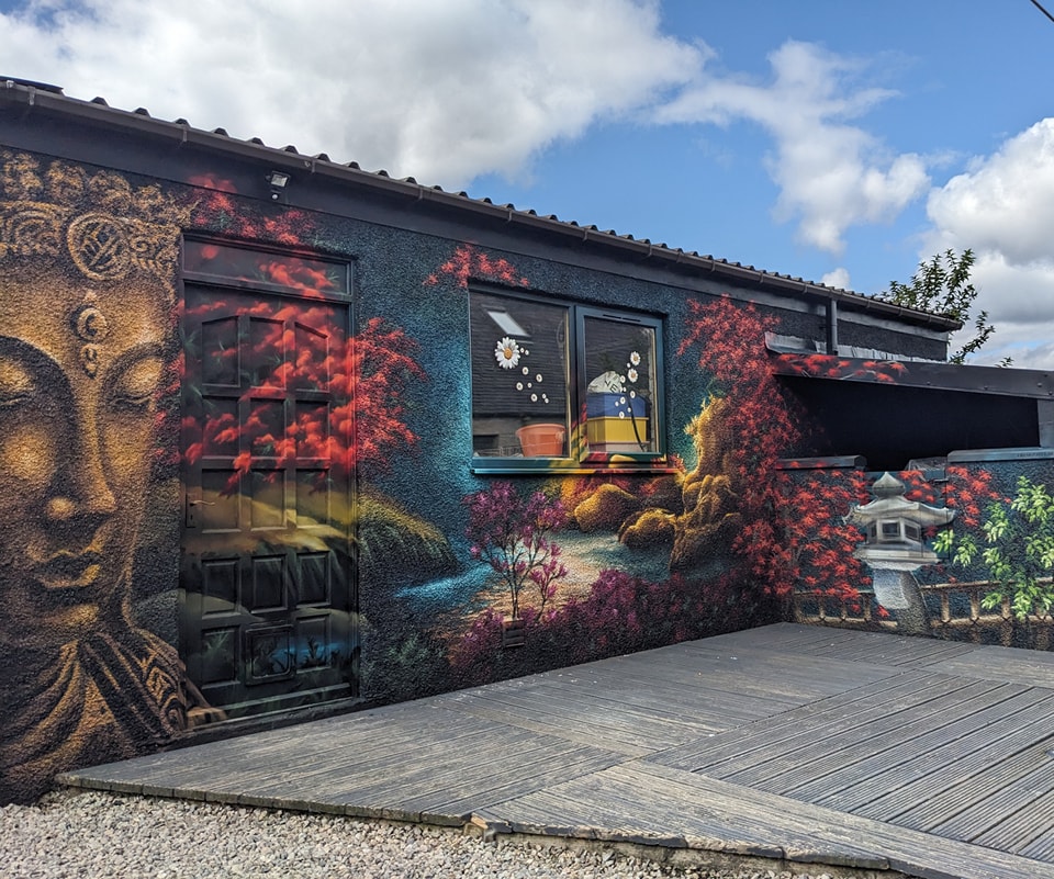 Mural featuring a large bronze Buddha face next to a doorway, surrounded by colorful Japanese garden scenery, a window, and a stone lantern, set against a blue, cloudy sky.