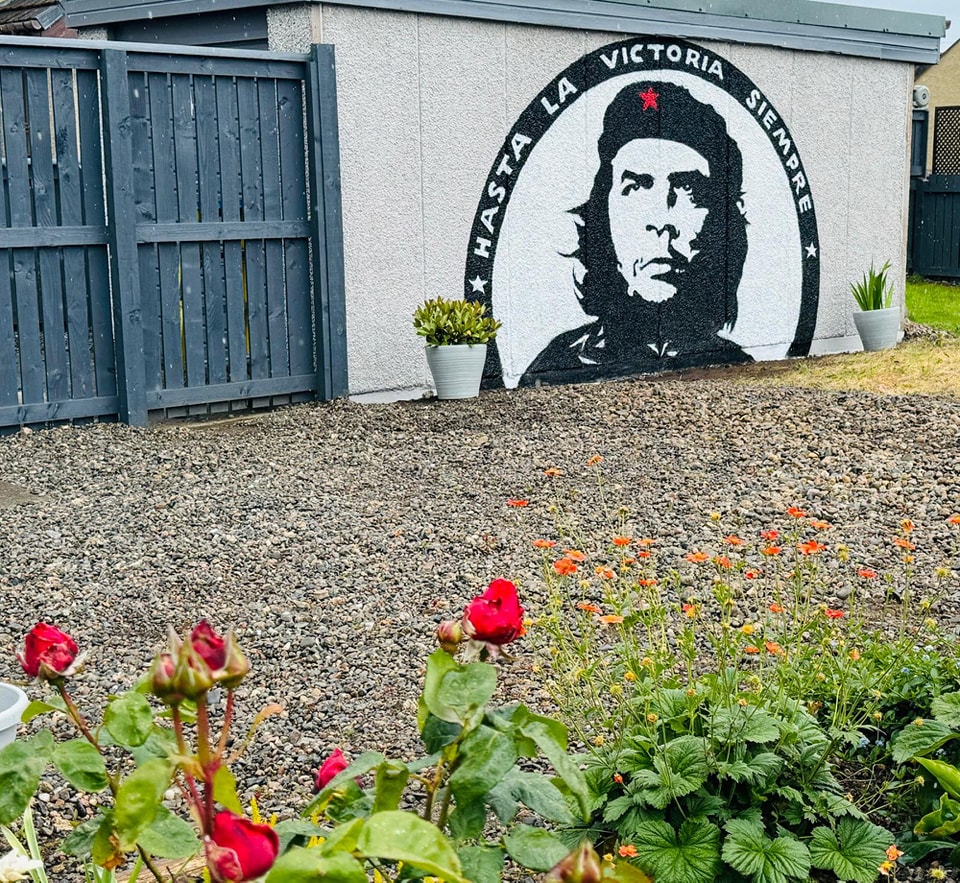 A black and white mural of Che Guevara with the text "Hasta la Victoria Siempre" is painted on a white shed next to a dark fence, overlooking a gravel yard with red roses and small orange flowers in the foreground.