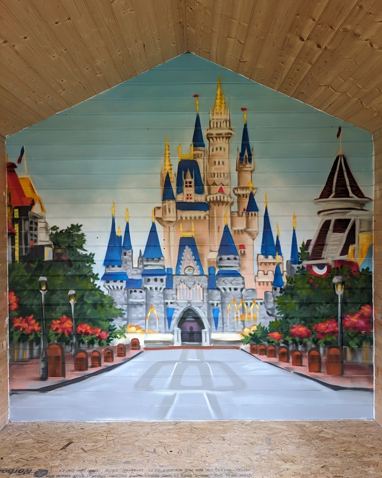 Mural painting of a fairytale castle with blue spires opening onto a path, framed by light wood paneling.