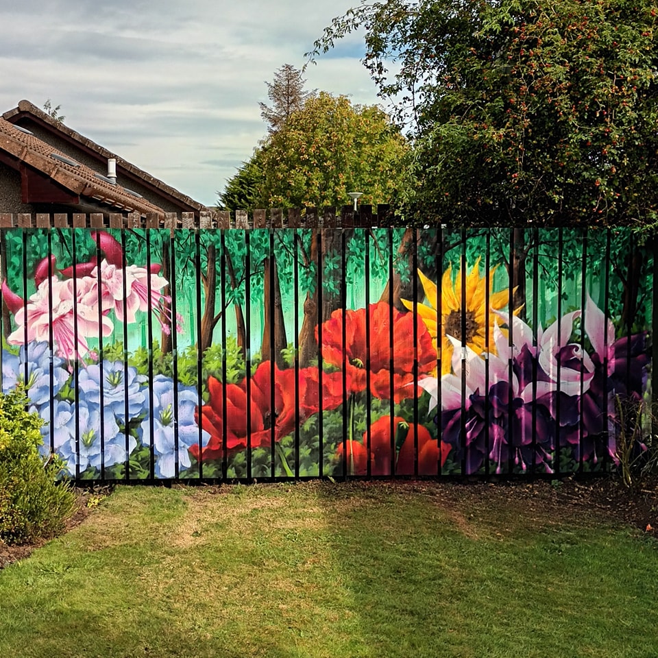 A brightly colored mural of large flowers and a forest scene painted across a vertical wooden garden fence overlooking a grassy lawn.