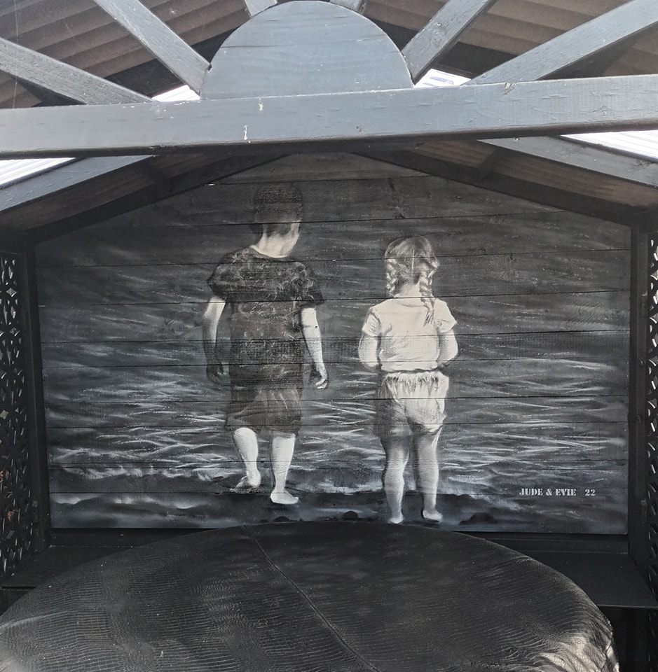 Black and white mural of a boy and girl standing at the water's edge, painted on wooden panels beneath a dark wooden roof structure.