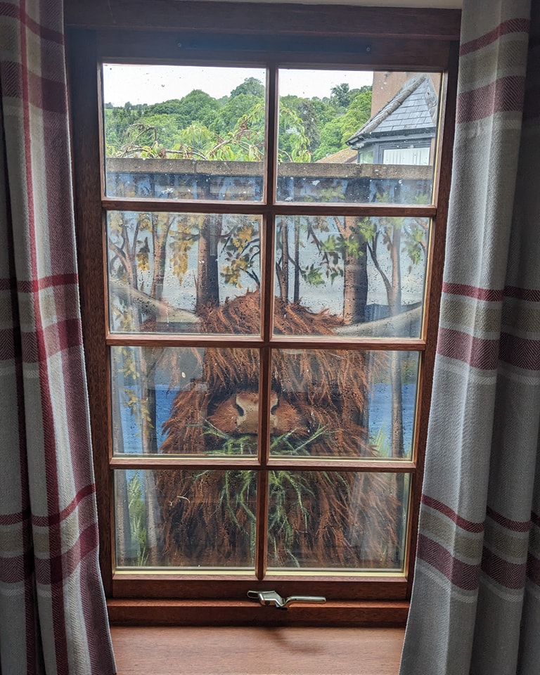 A window with a four-pane painting of a shaggy, brown Highland cow eating grass is framed by plaid curtains, overlooking green hills outside.