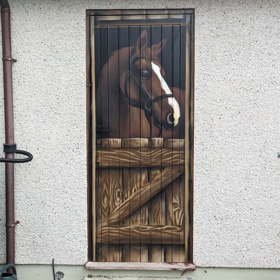 Trompe l'oeil door painted to look like a brown horse with a white blaze peering over the top half of a wooden stable door mounted on a white stucco exterior wall.