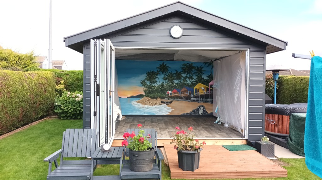 A modern grey garden shed with bi-fold doors open reveals an interior mural of a tropical sunset beach scene, with patio furniture and potted flowers outside.