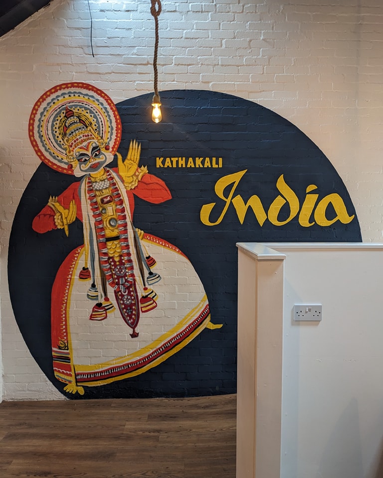 Mural featuring a colorful Kathakali dancer painted on a dark circle against a white brick wall