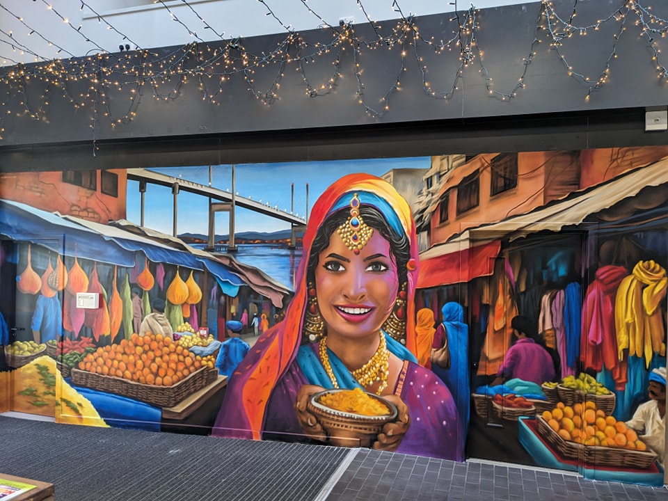 Vibrant mural located in the Victorian Market in Inverness depicting a smiling South indian woman holding spices in a busy outdoor market scene with the North Kessock bridge in the background.