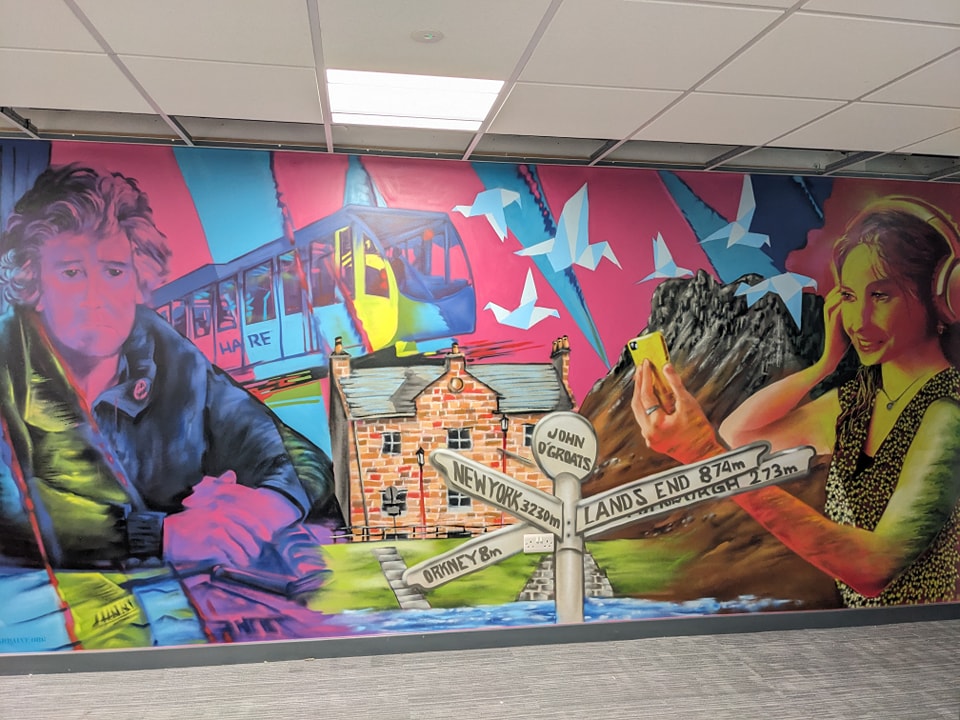 Vibrant mural painted at the Inverness Courrier office featuring portraits of two individuals, Eveline Barron – who held sway over the content of the Inverness Courier for 70 years until 1988 – and Iona MacDonald, Highland News and Media's (HNM) youngest reporter who, is already producing important in-depth stories for both print editions and online. a signpost indicating distances to locations like New York, John O'Groats, and Lands End, against a pink background with a cable car from the Cairngorms and stylized paper birds.