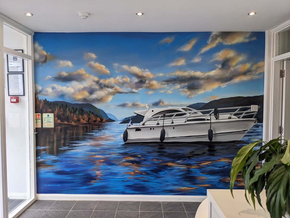 Mural depicting a yacht on deep blue Loch Ness water against a backdrop of forested mountains under a dramatic, cloudy sky.
