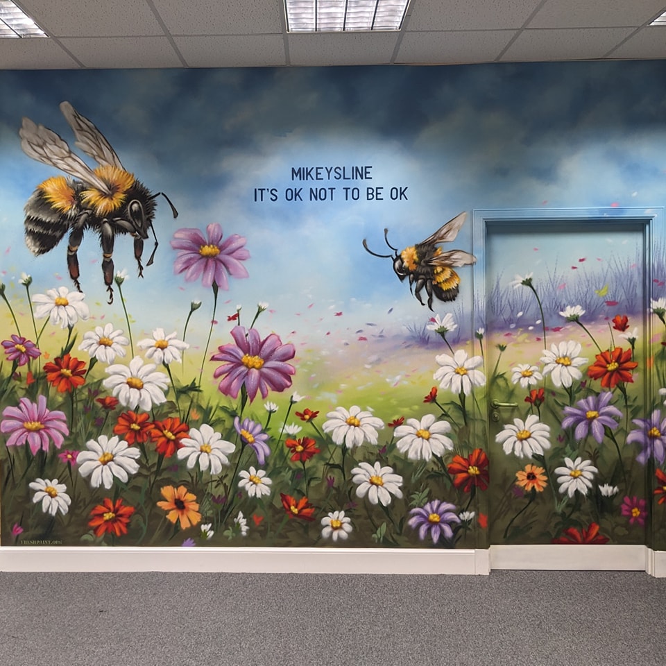 A colorful mural featuring two large bumblebees flying over a field of daisies and purple flowers, with the text "MIKEYS LINE IT'S OK NOT TO BE OK" painted on a blue, cloudy sky background.