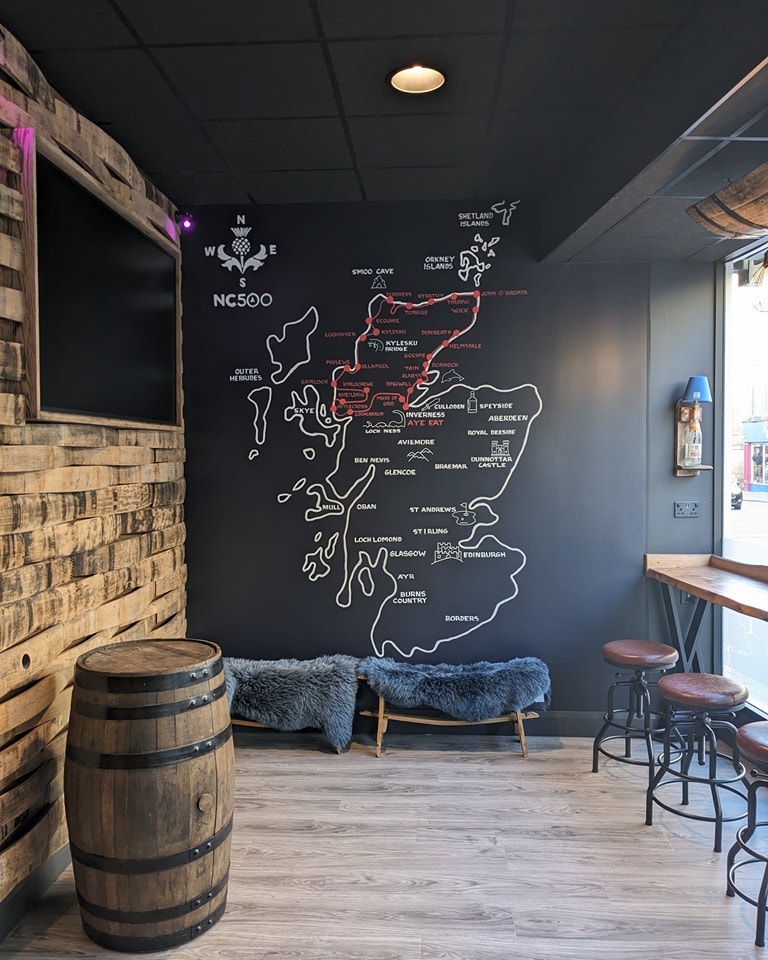 Interior view of a modern bar, restaurant Aye Eat in Inverness featuring a large black wall with a white chalk-style map of the North Coast 500 (NC500) route in Scotland, a wooden whiskey barrel, and leather bar stools.