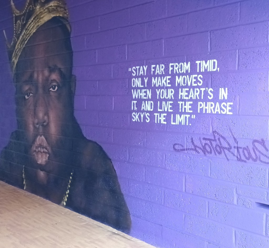 Mural of The Notorious B.I.G. wearing a crown next to a white stencil quote reading, "Stay far from timid, only make moves when your heart's in it, and live the phrase sky's the limit," on a purple brick wall.