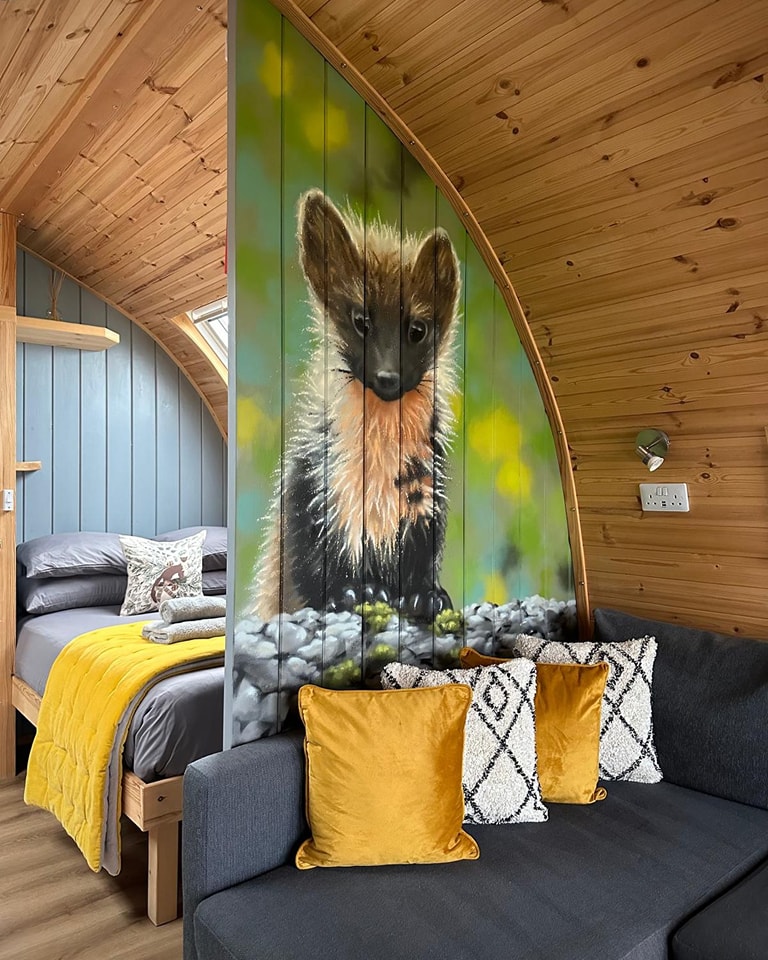Interior view of a cozy wooden cabin featuring a prominent mural of a small brown pine marten dividing a sleeping area with a yellow-accented bed and a grey sofa with mustard throw pillows.