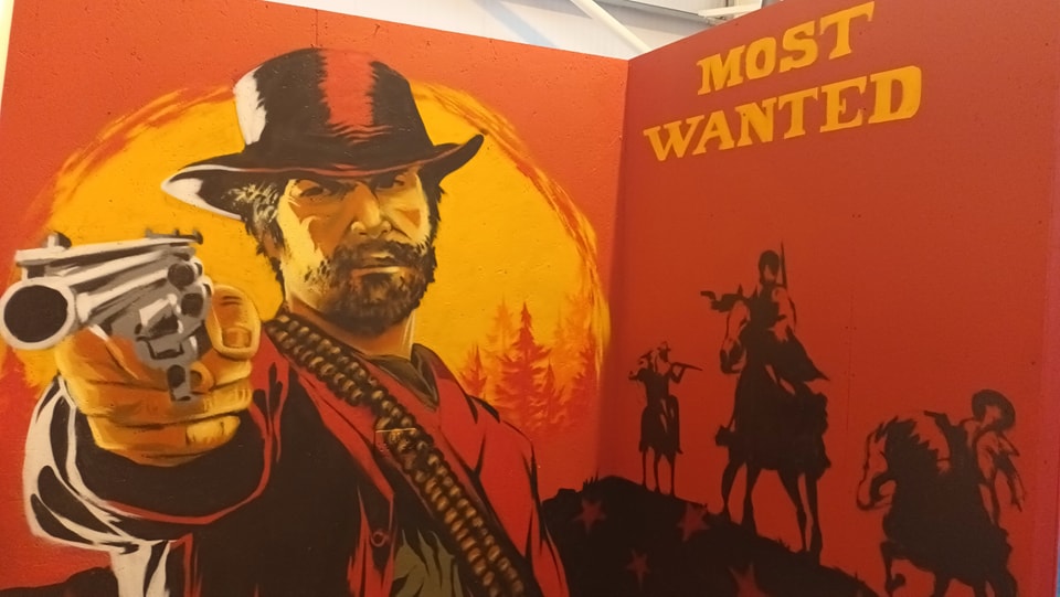 ainted promotional art Red Dead Redemption, showing a bearded cowboy pointing a revolver forward, set against an orange and red background with "MOST WANTED" text and silhouetted figures on horseback.