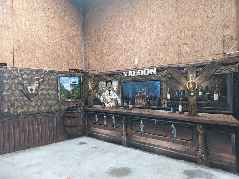 Mural painting depicting a rustic Western saloon bar with a bartender, mounted stag head, and whiskey barrel against a raw wood-paneled wall.