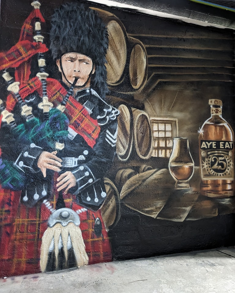 Mural painting featuring a bagpiper in traditional Scottish attire next to whisky barrels, a dram glass, and a bottle labeled "Aye Eat 25."
