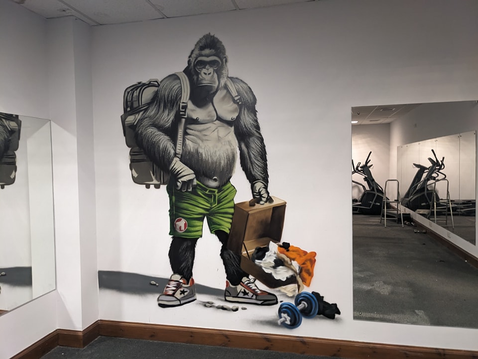 A large wall mural depicts a muscular silverback gorilla wearing a backpack, green shorts, and sneakers, unpacking a suitcase next to a dumbbell in a gym with mirrored walls reflecting exercise equipment.
