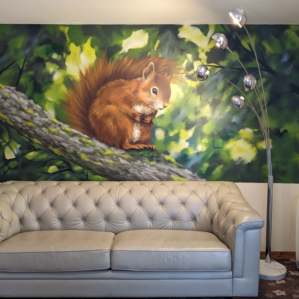 A light gray tufted leather sofa sits beneath a large, colorful wall mural depicting a red squirrel perched on a branch amidst green foliage, next to a modern chrome floor lamp.