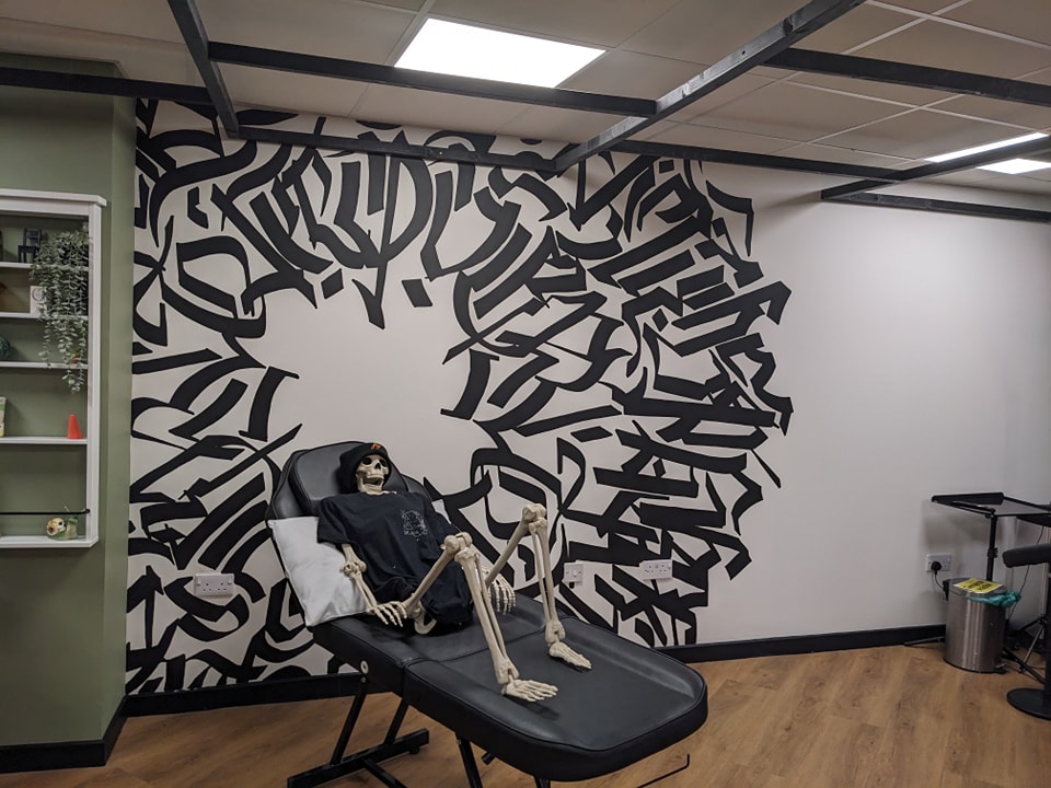 A skeleton wearing a black hoodie sits on a black tattoo armchair against a white wall featuring a large black abstract calligraphy mural at ERA tattoo Inverness.