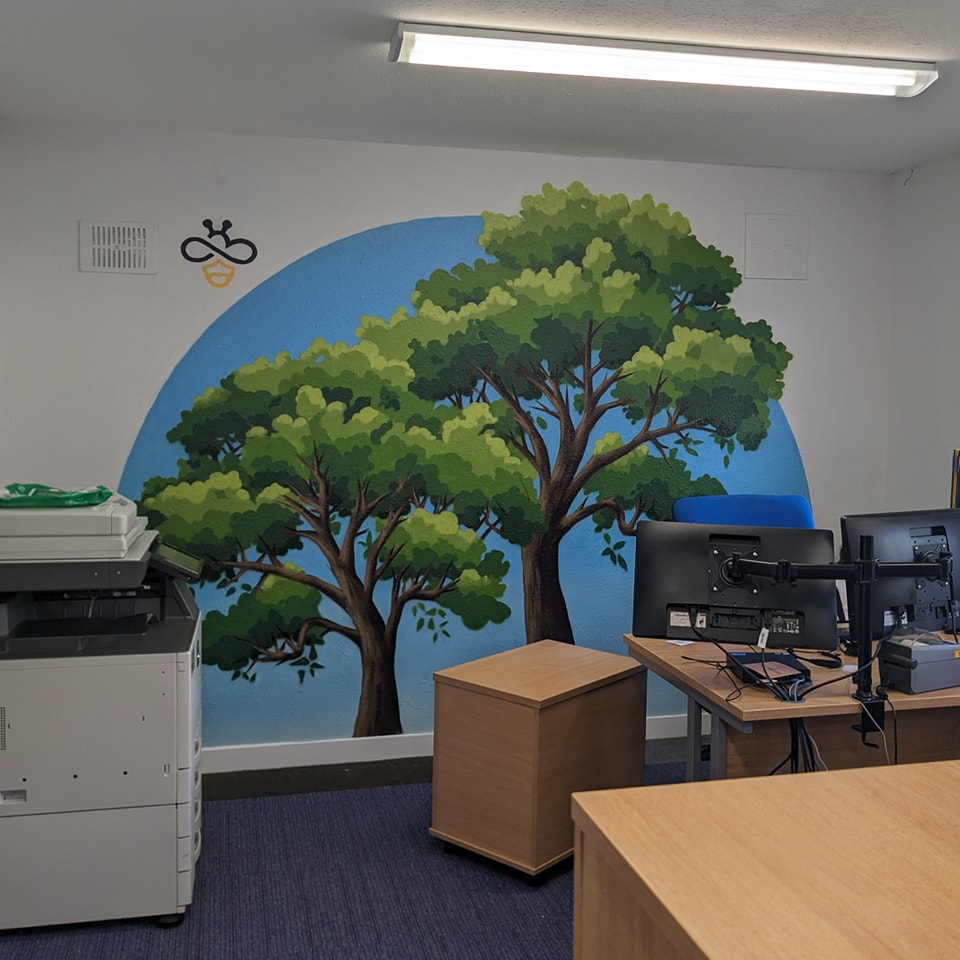 Office interior featuring a large wall mural of two green trees against a blue sky circle, near a copier and computer workstations.