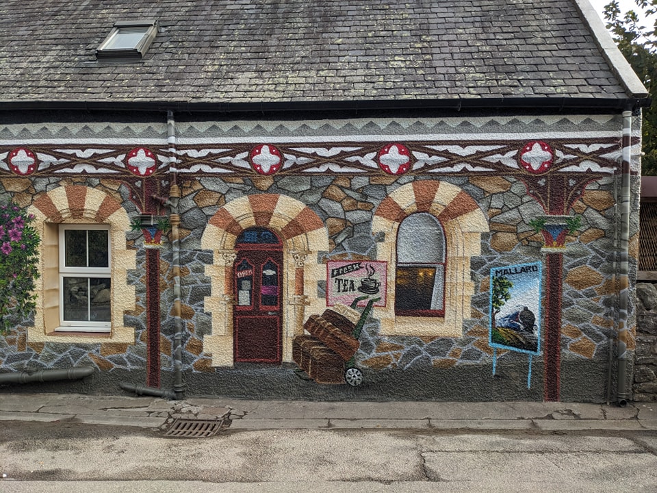 Mural painting in Drumnadroquit of a Victorian inspired train station with a stone building facade depicting arched windows, a dark red door, and signs for "Fresh Tea" and the "Mallard" train.