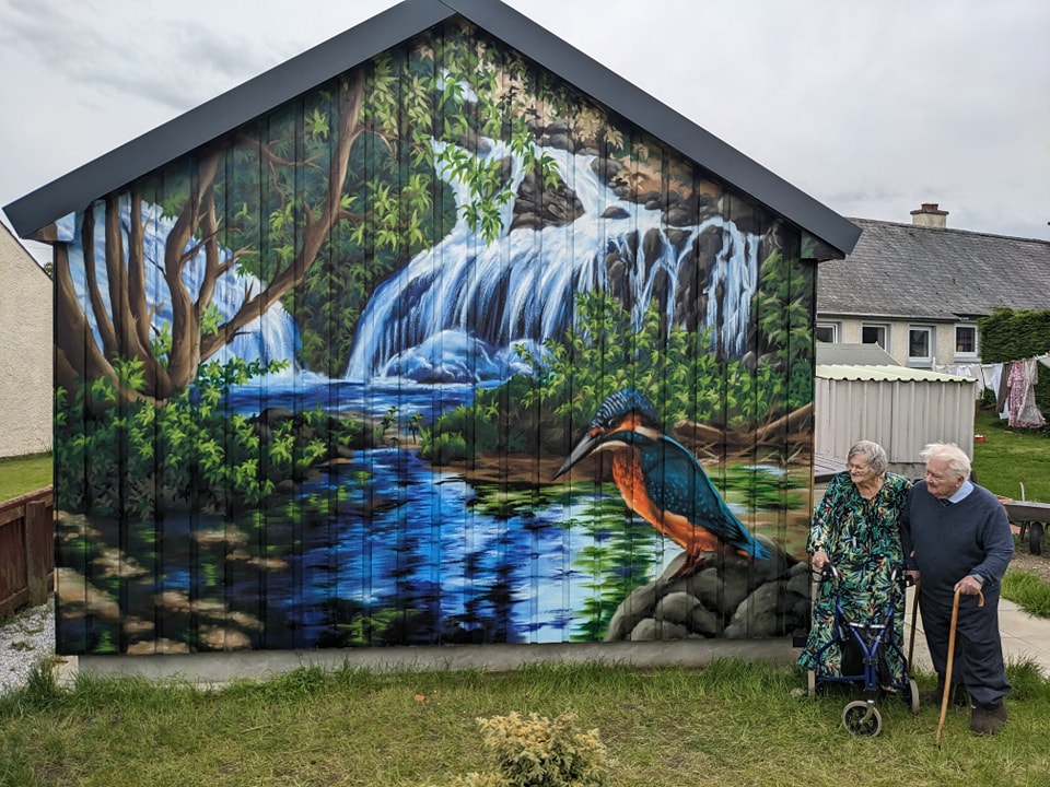 An elderly couple stands next to a garden shed painted with a vibrant mural of a woodland waterfall scene featuring a large kingfisher perched on rocks above a blue stream.