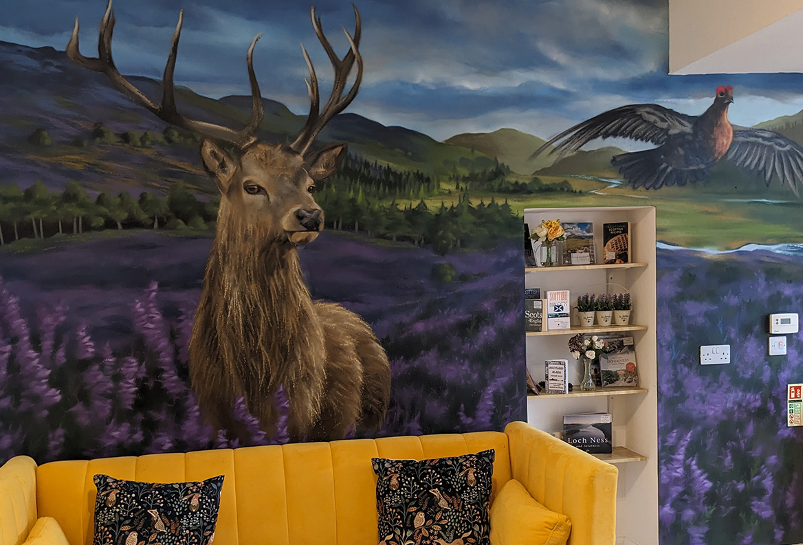 A vibrant room in a airbnb featuring a large Scottish-themed mural of a stag in purple heather, a flying grouse, and rolling hills above a bright yellow sofa with patterned cushions, next to built-in shelving with local Highlands info.