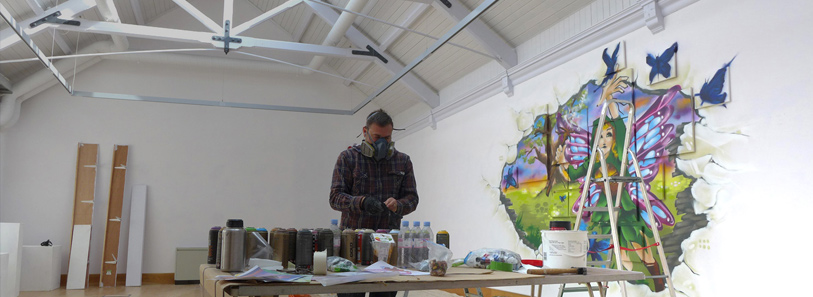 A graffiti artist wearing a respirator stands in a bright studio setting surrounded by spray cans while working on a vibrant fairy mural on the white wall.