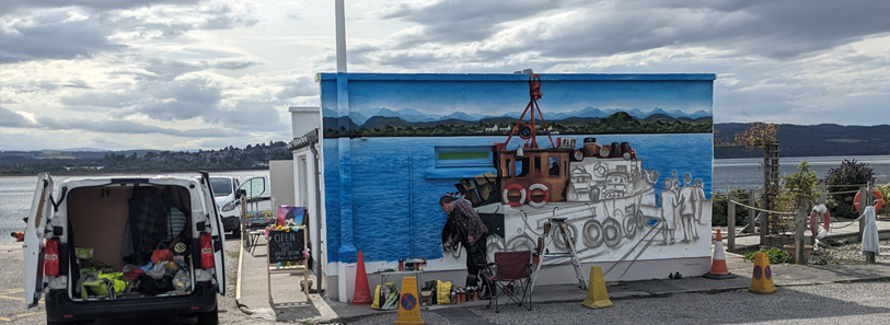 Mural Artist at work, with a partially painted boat and figures against a blue sea and mountain backdrop is being worked on outdoors next to a lamppost and Fresh Paint white van.
