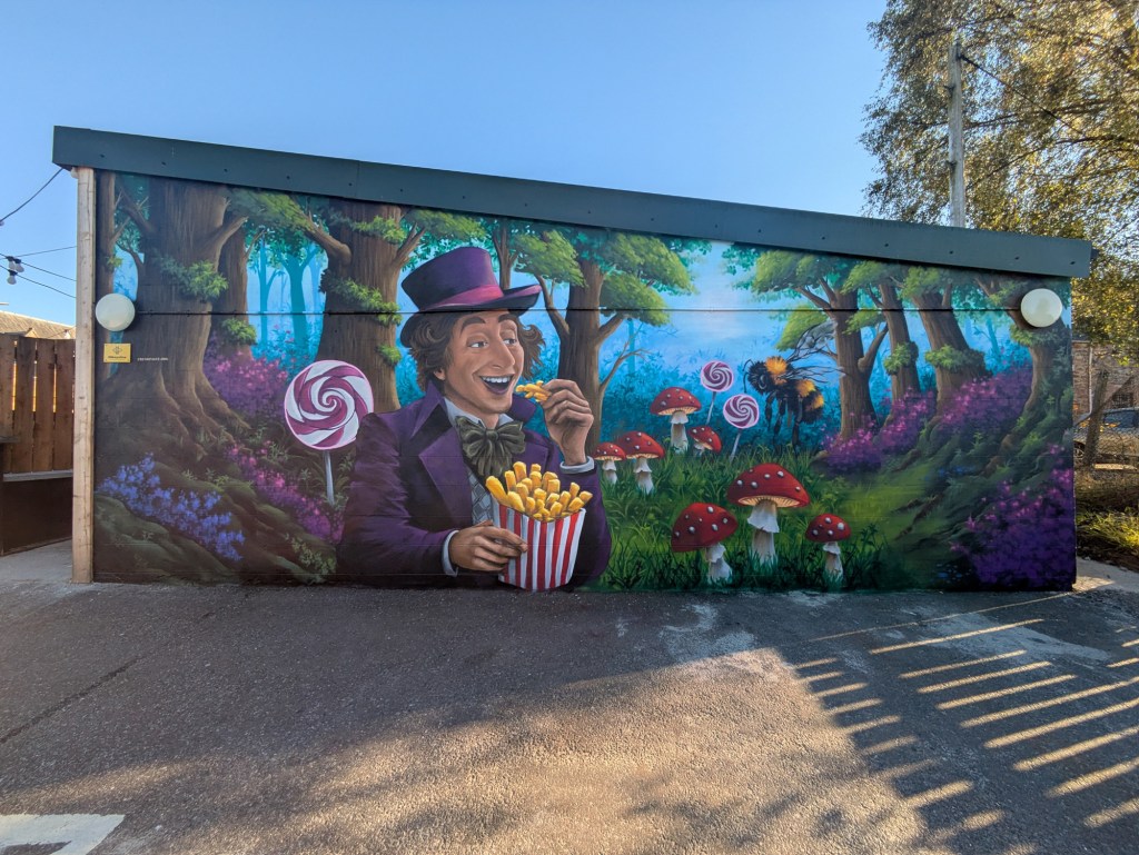 Willy Wonka mural painted on the Tarradale chippy wall in the Muir of Ord, featuring him eating chips amidst a colorful forest setting with toadstools, lollipops and a large Mikeysline bee.